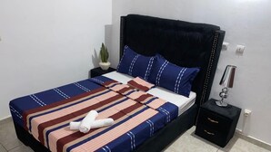 Una cama bien hecha con ropa de cama a rayas, una toalla enrollada, una mesita de noche con una lámpara y una planta en maceta.