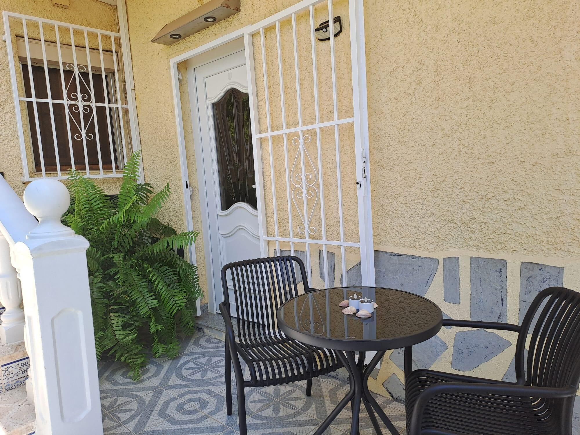 An outdoor seating area with a round table and two chairs, a white door with a decorative design, and a white railing.