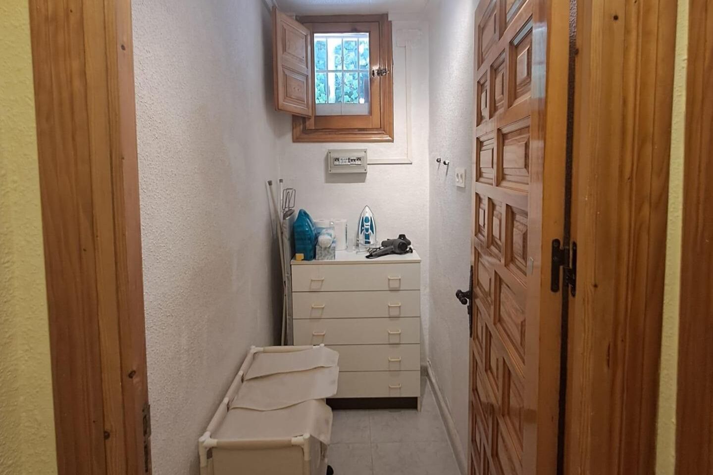 Un couloir étroit, avec une porte en bois sur la droite, une fenêtre donnant sur un paysage verdoyant et une petite armoire blanche sur laquelle sont posés divers objets.