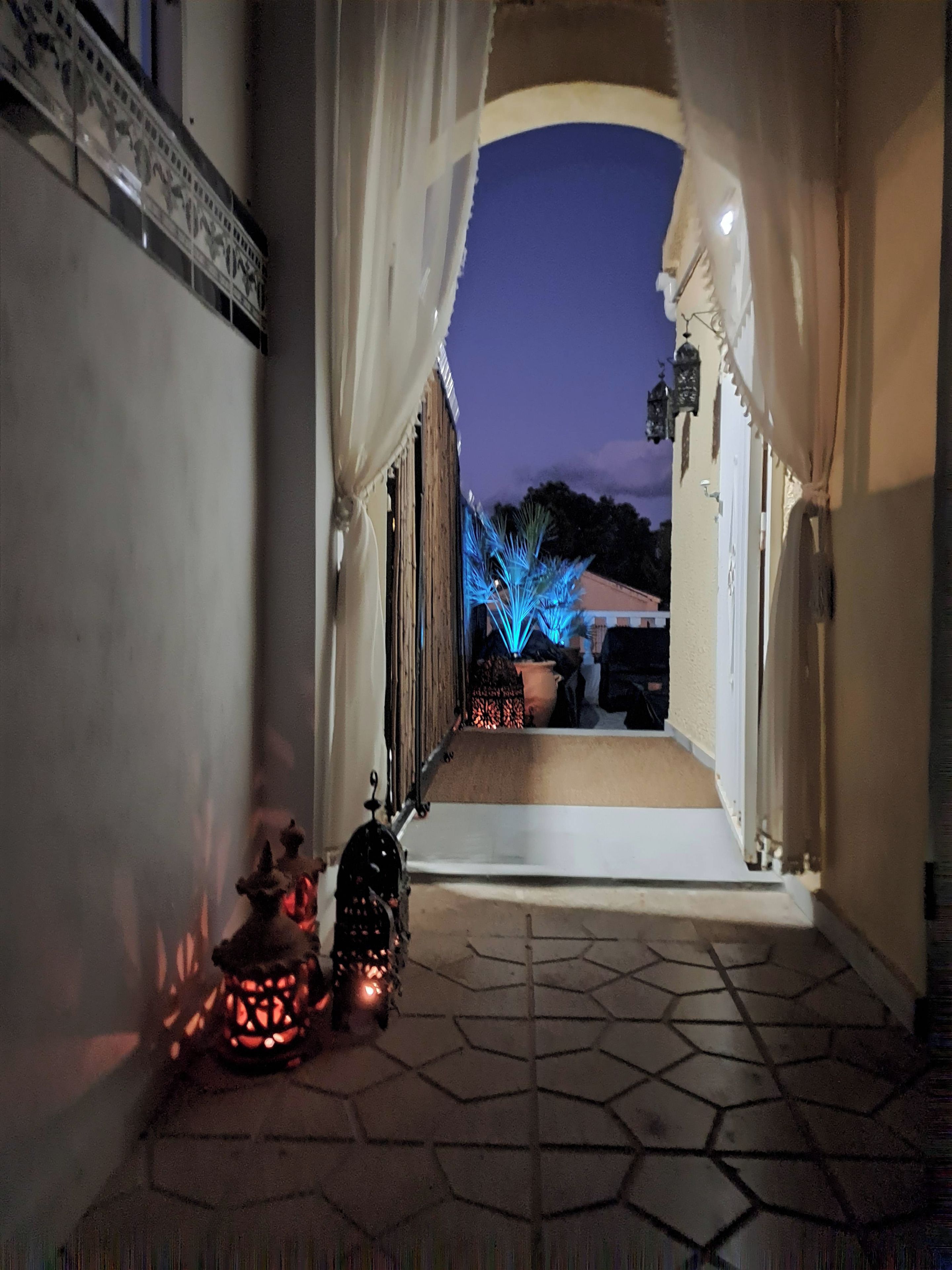A corridor with a tiled floor leading to an outdoor area with a potted plant and a decorative light.
