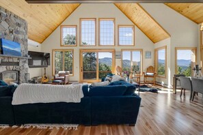 A spacious living room with a stone fireplace, a large blue sofa, and multiple windows offering a view of the mountains.