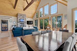 A spacious living room with a large wooden table, a stone fireplace, and a television mounted on the wall.