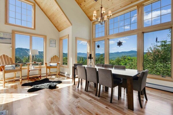 A dining area with a large table and chairs, large windows with a mountain view, and a wooden ceiling.