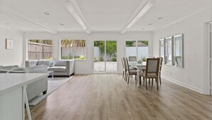 A spacious room with a dining area, a sofa, and a view through large windows. - Driftwood Cottage | Luxury 3BR Pool Home Steps to East Beach St. Simons Island (Saint Simons Island)