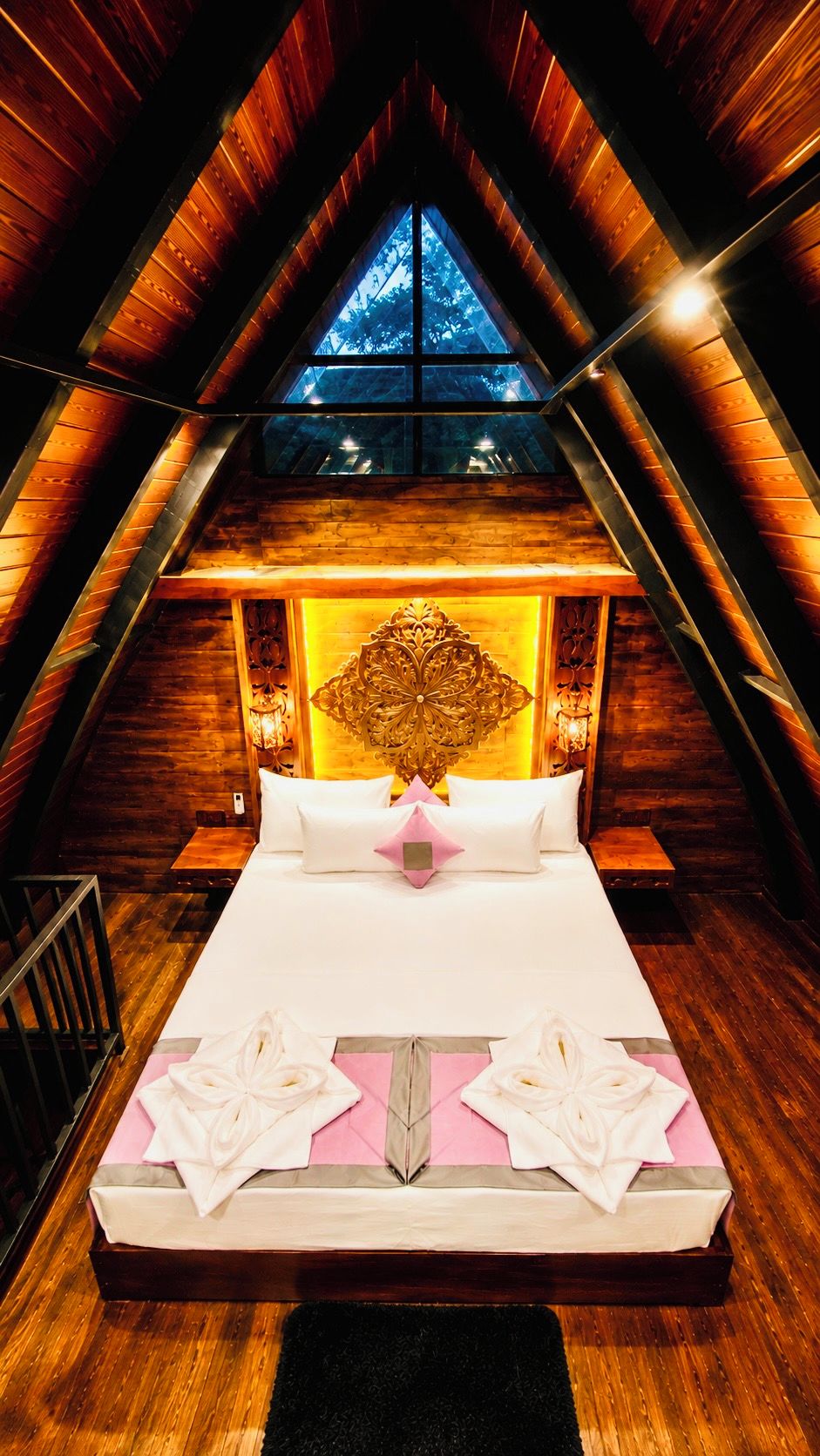 A high-ceilinged room with a wooden sloped roof, a large bed with white linens, and a decorative headboard.