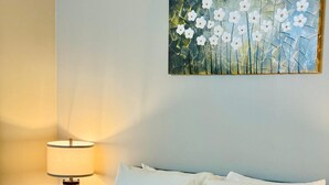 Un lit bien fait, avec une literie blanche et un plaid rayé jaune et blanc. Un tableau représentant des fleurs blanches est accroché au mur, au-dessus du lit.