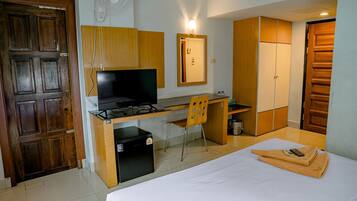 Habitación de hotel pequeña con cama, escritorio con televisor, refrigerador compacto, una silla y armarios de madera.