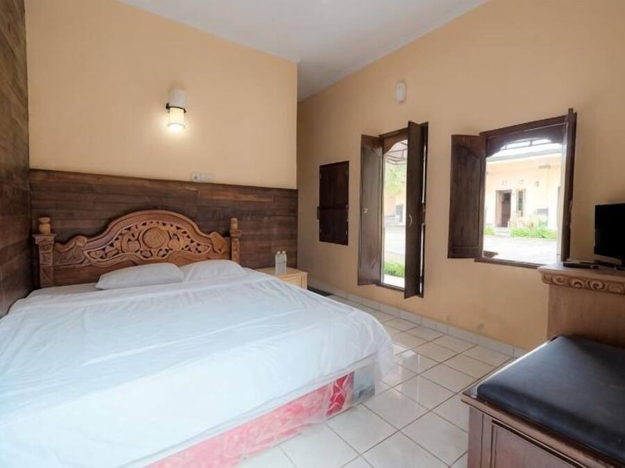 Un dormitorio con cabecera de madera, una cama, una mesita de noche, un banco, un televisor y una ventana con puerta abierta.