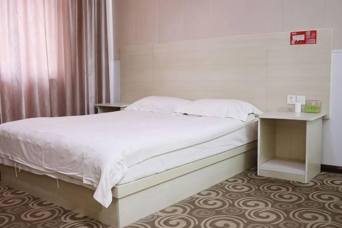Quarto de hotel com uma cama, uma mesinha de cabeceira, um interruptor de luz fixado na parede e um tapete estampado.