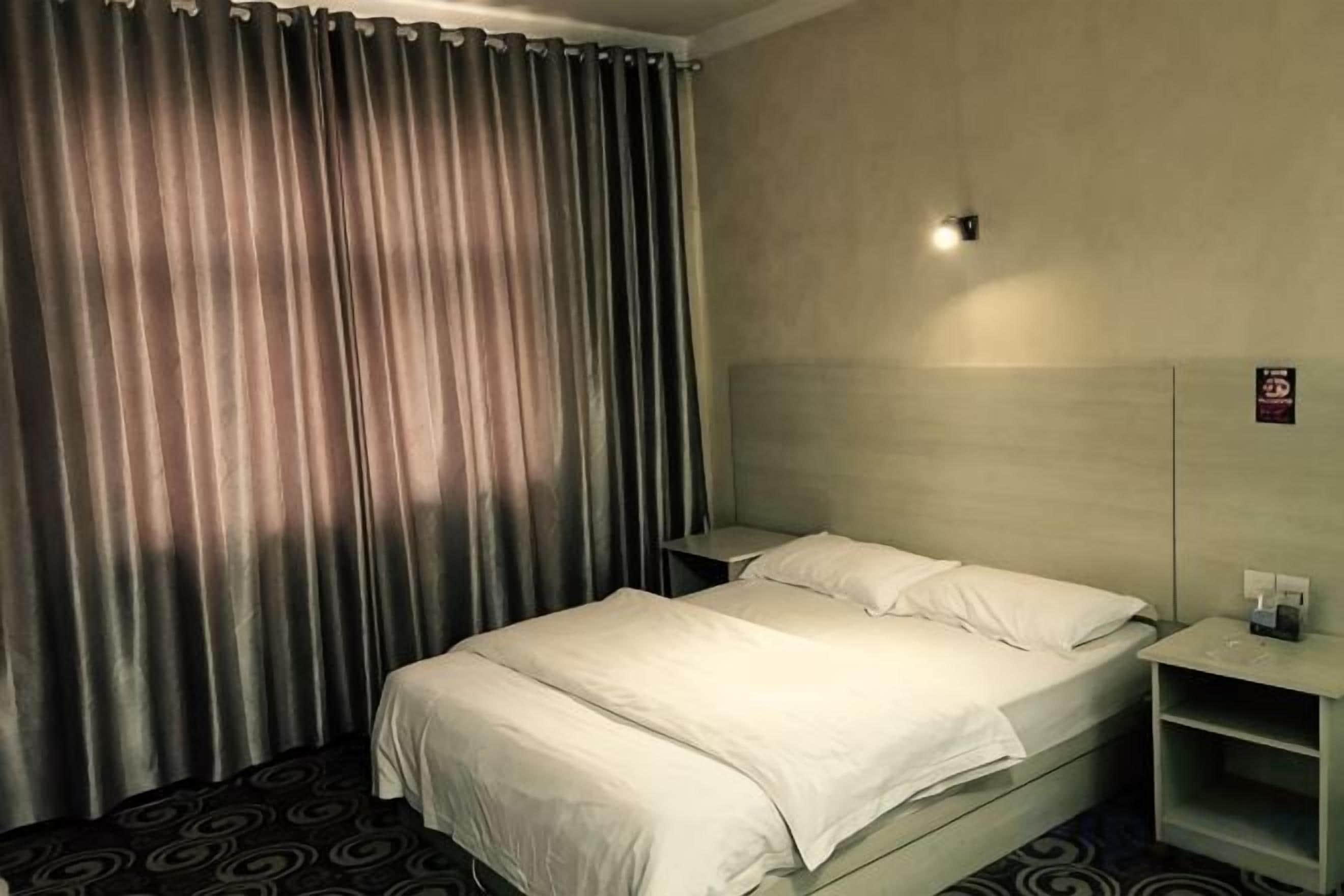 A hotel room with a bed, curtains, a nightstand, and a wall-mounted light.