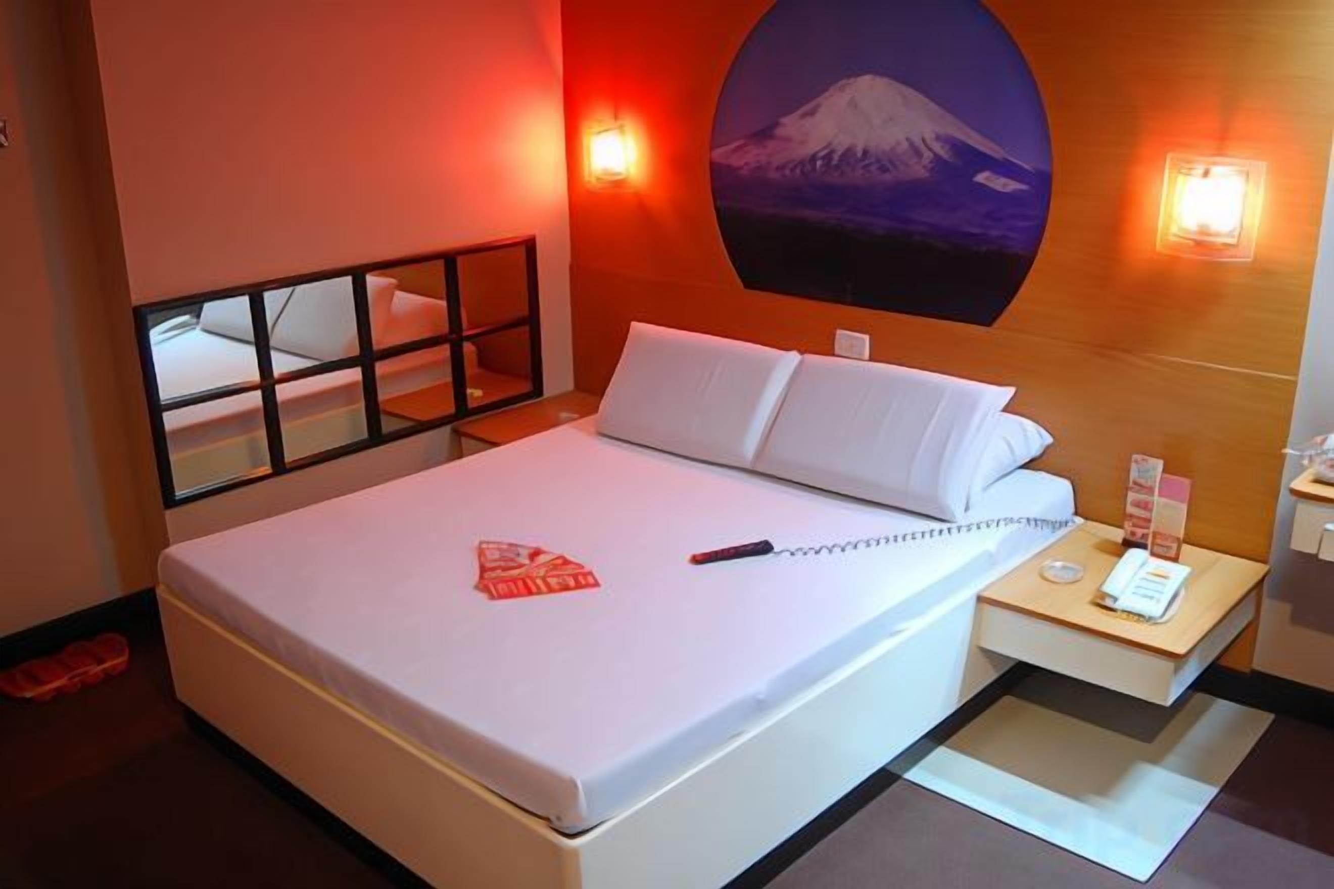 A hotel room with a bed, a bedside table with a lamp, a wall-mounted TV, and a picture of Mount Fuji.