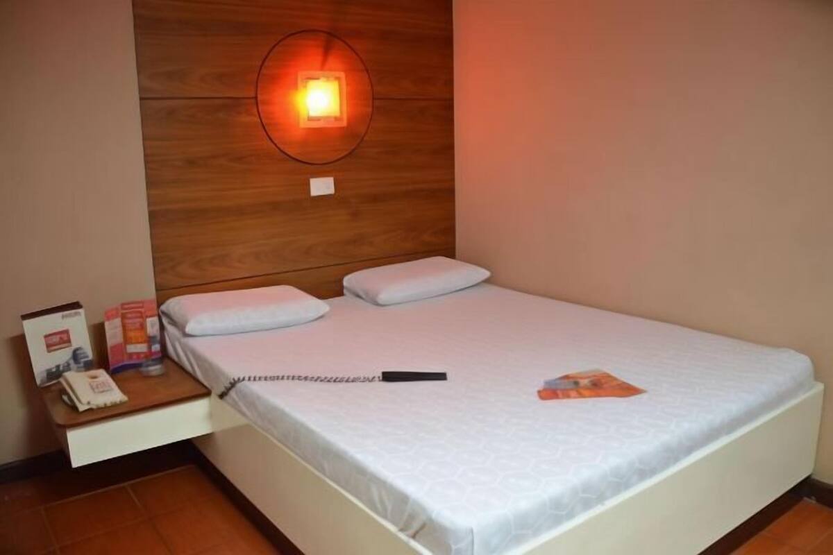 Una cama individual con colchón y almohadas blancos, una mesita de noche con teléfono y artículos de aseo, y una lámpara fijada a la pared.