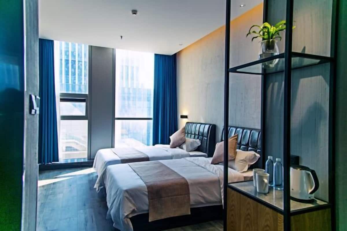 Moderni hotellihuone, jossa on kaksi s änkyä, suuri ikkuna sinisillä verhoilla, yöpöytä, jossa on vedenkeitin, ja hyllyllä oleva kasvi.