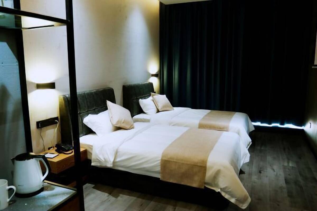 Hotellihuone, jossa on kaksi sänkyä, yöpöytä lampun kanssa, kahvinkeitin ja verho.