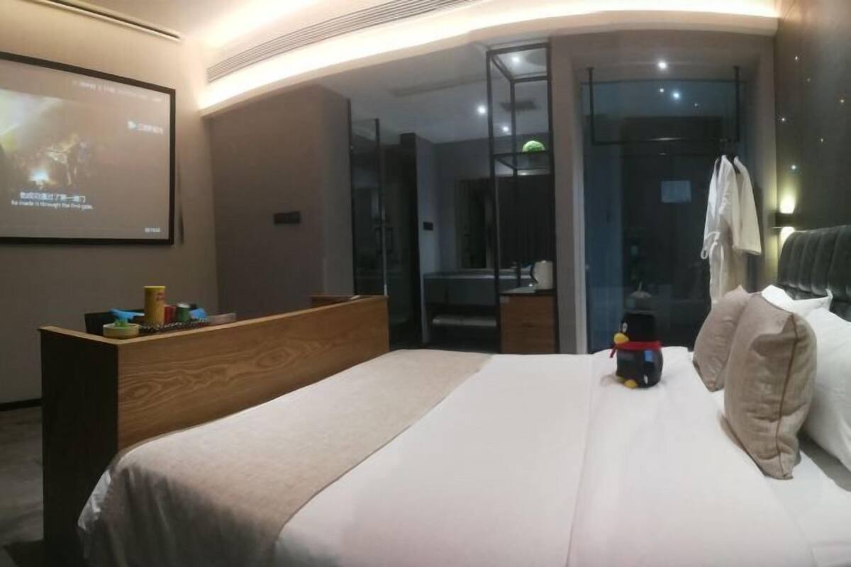 Moderni hotellihuone, jossa on suuri sänky, puinen työpöytä, lasinen suihkukaappi ja seinälle asennettu projektorikangas.