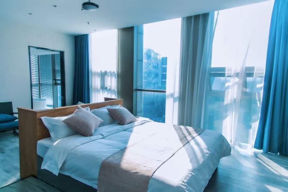 Moderni hotellihuone, jossa on suuri sänky, yöpöytä, peili ja suuret ikkunat, joista avautuu näkymä kaupunkiin.