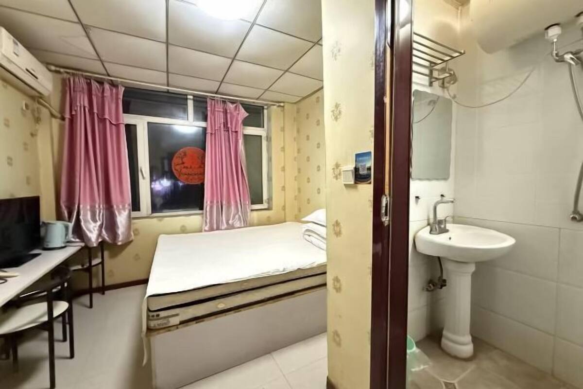 A small room with a bed, a desk, a window with curtains, a sink, and a shower.