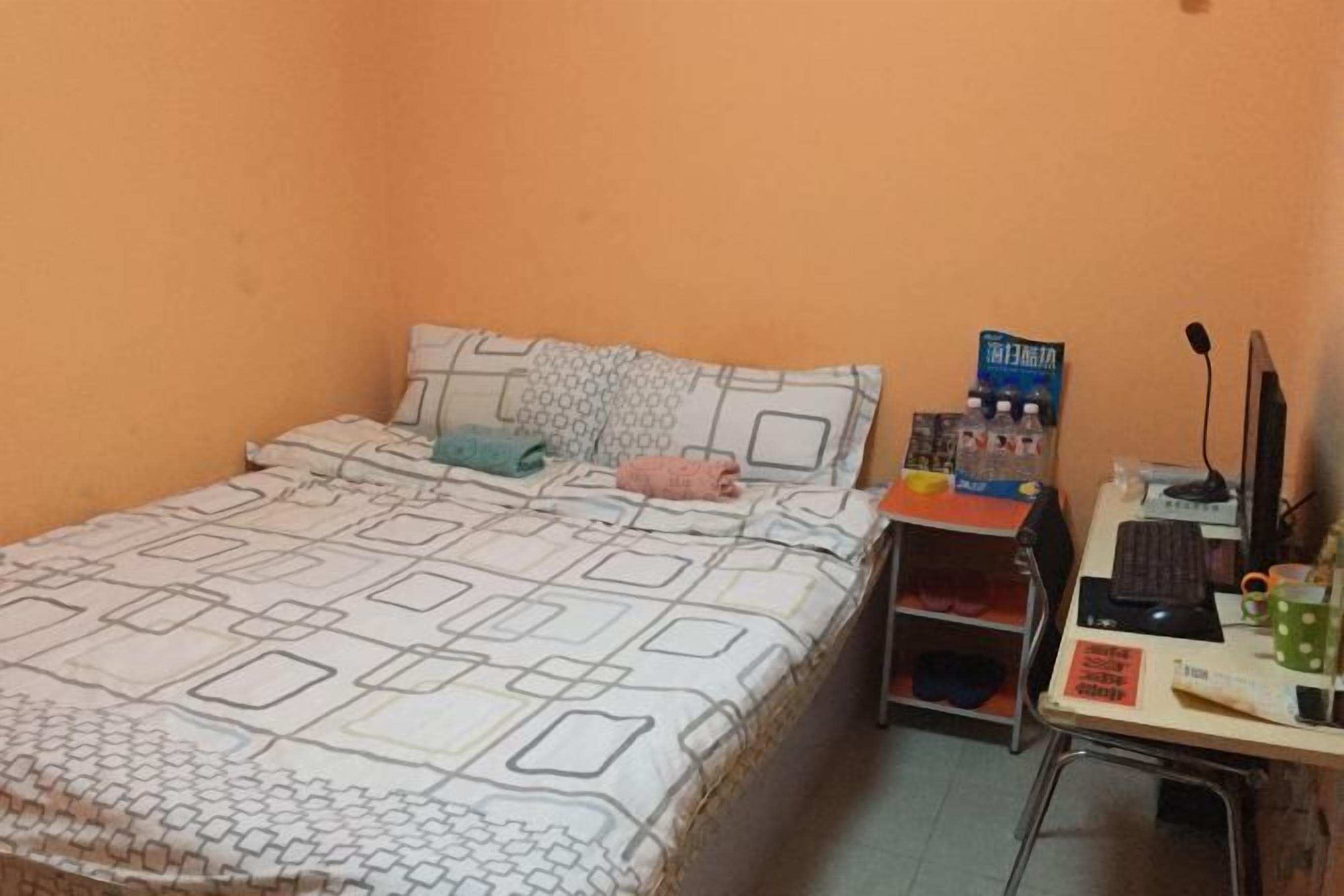 A bedroom with a bed, a desk with a computer, a chair, and a shelf with items.