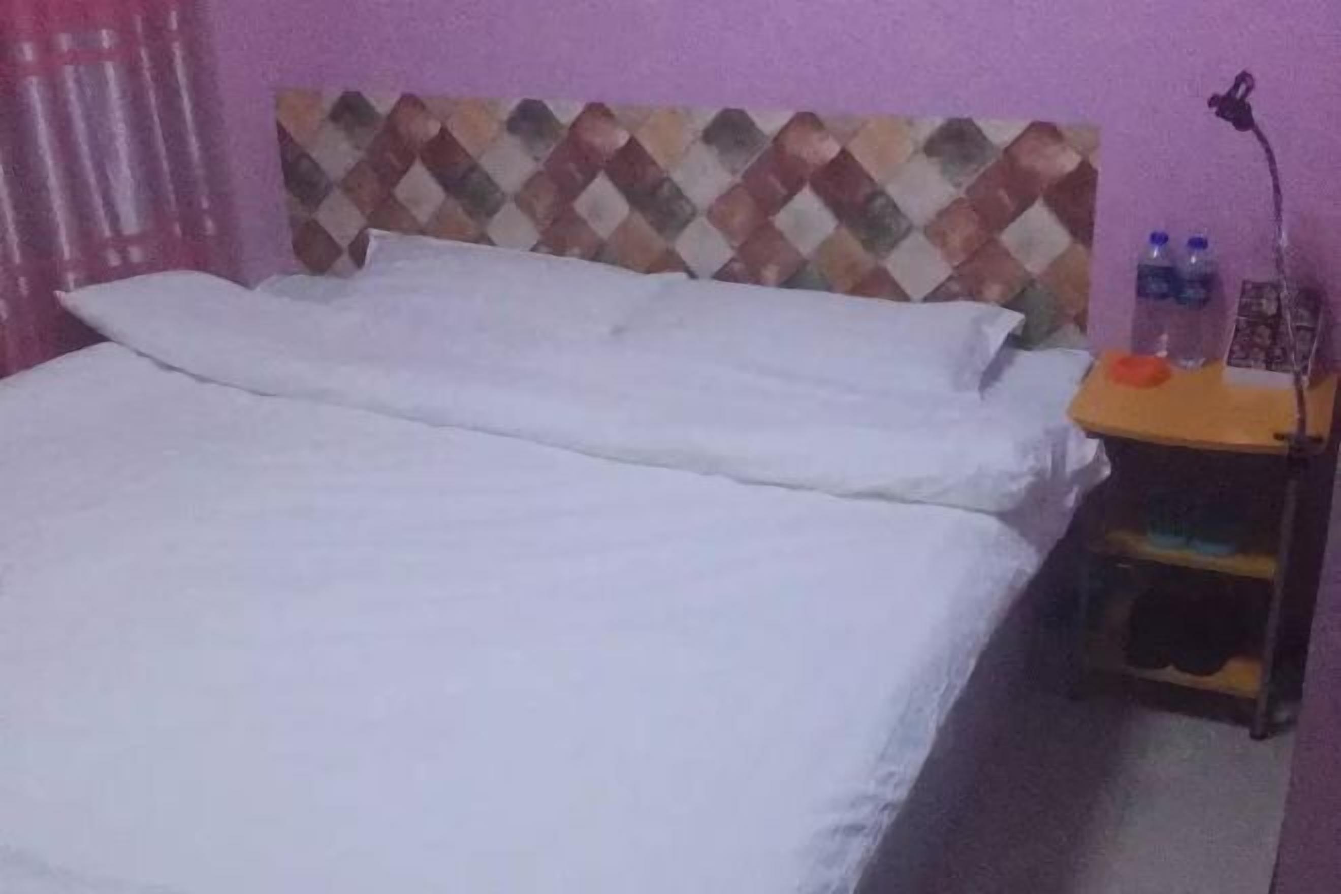 A bed with white bedding, a headboard with a diamond pattern, a nightstand with a lamp, and two water bottles.