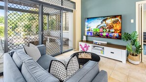 Ein modernes Wohnzimmer mit einem grauen Sofa, einem Fernseher auf einer weißen Unterhaltungsanlage und einer Glastür, die zu einem Außenbereich führt.