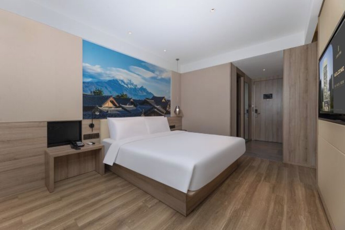 Une chambre d’hôtel moderne avec un grand lit, un téléviseur à écran plat, une table de chevet en bois et un tableau représentant des montagnes accroché au mur.