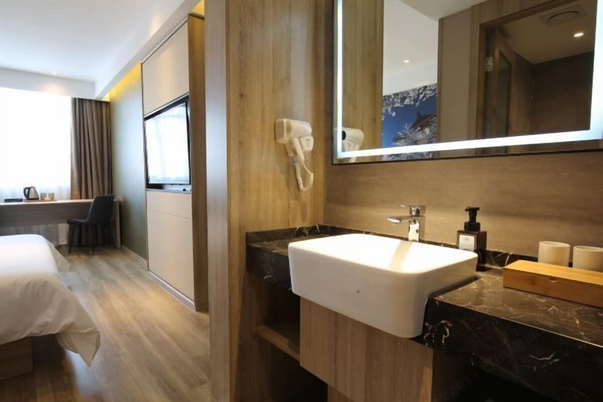 Une chambre d’hôtel moderne avec un plancher en bois, un lit, un bureau avec une chaise, un grand miroir, un lavabo blanc et une commode avec des articles de toilette.