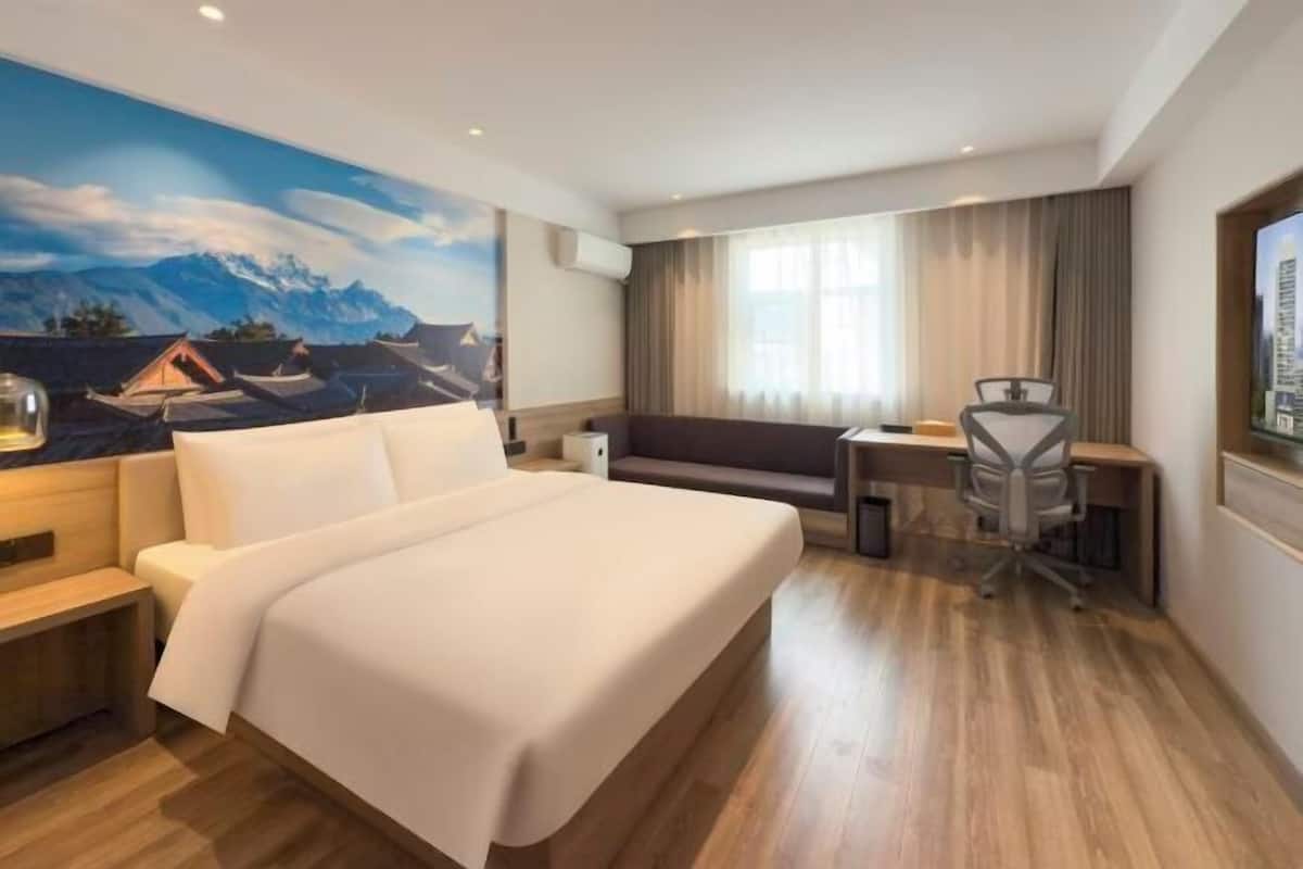 A hotel room with a large bed, a desk, a chair, a sofa, and a painting of mountains.