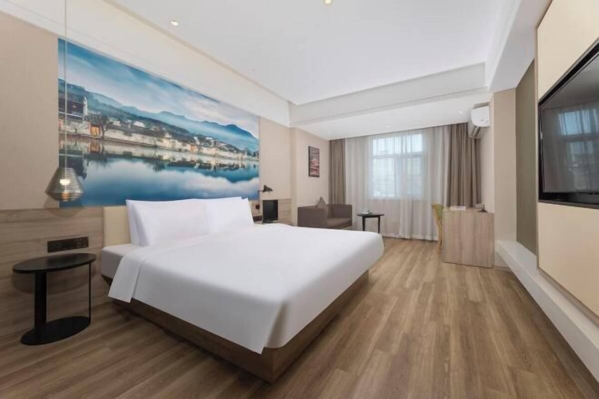 A hotel room with a large bed, a painting of a landscape, a TV, and a desk.