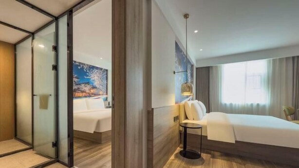 A hotel room with a bed, a bedside table, a lamp, a painting on the wall, and a sliding glass door leading to another room. - Yilong Anyun Hotel (Rudong Chahe Middle School) (Rudong)