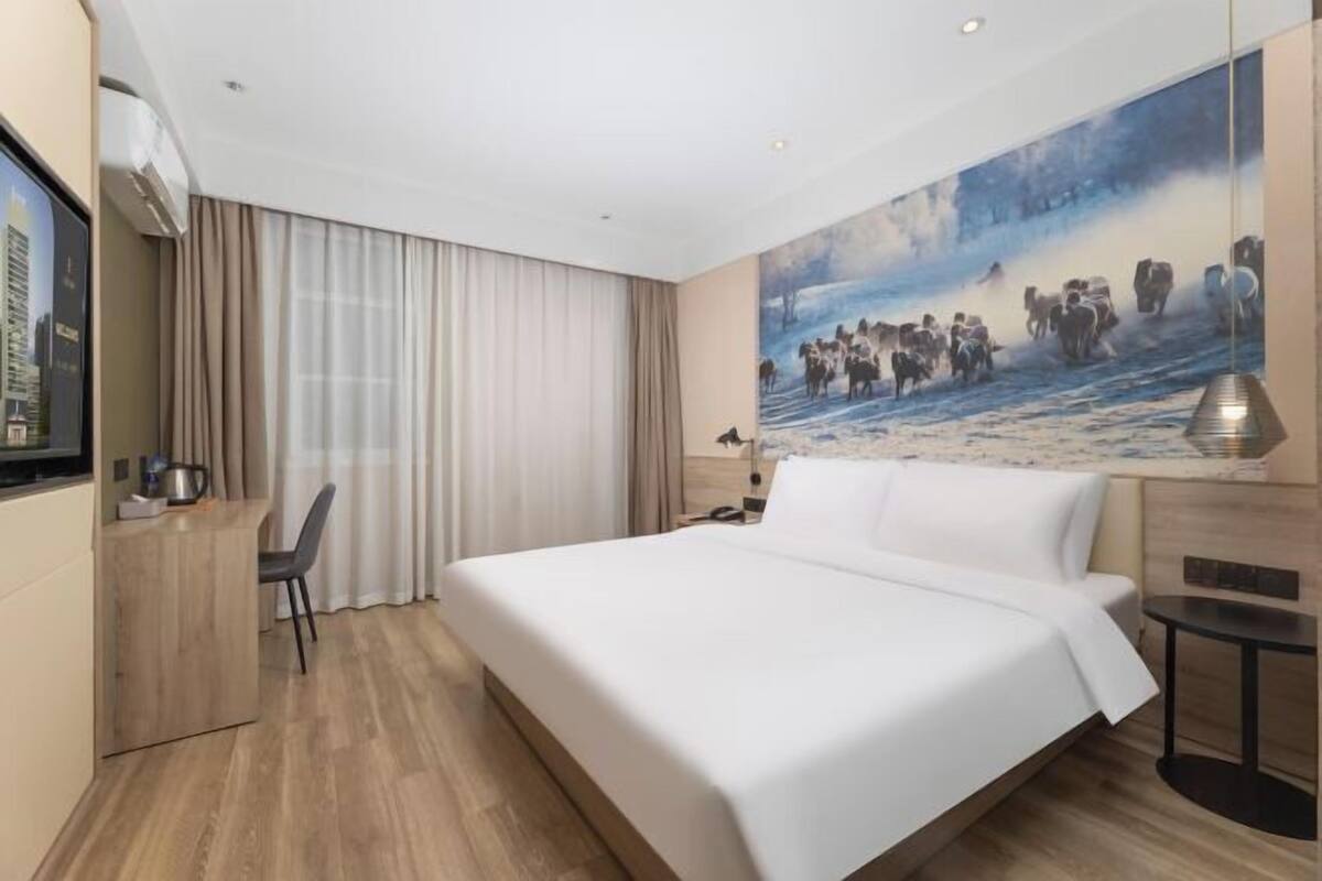 A hotel room with a large bed, a desk with a chair, a painting on the wall, and a window with curtains.