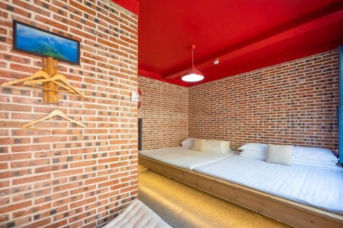A room with a brick wall, a bed, a hanging light fixture, and a television mounted on the wall.