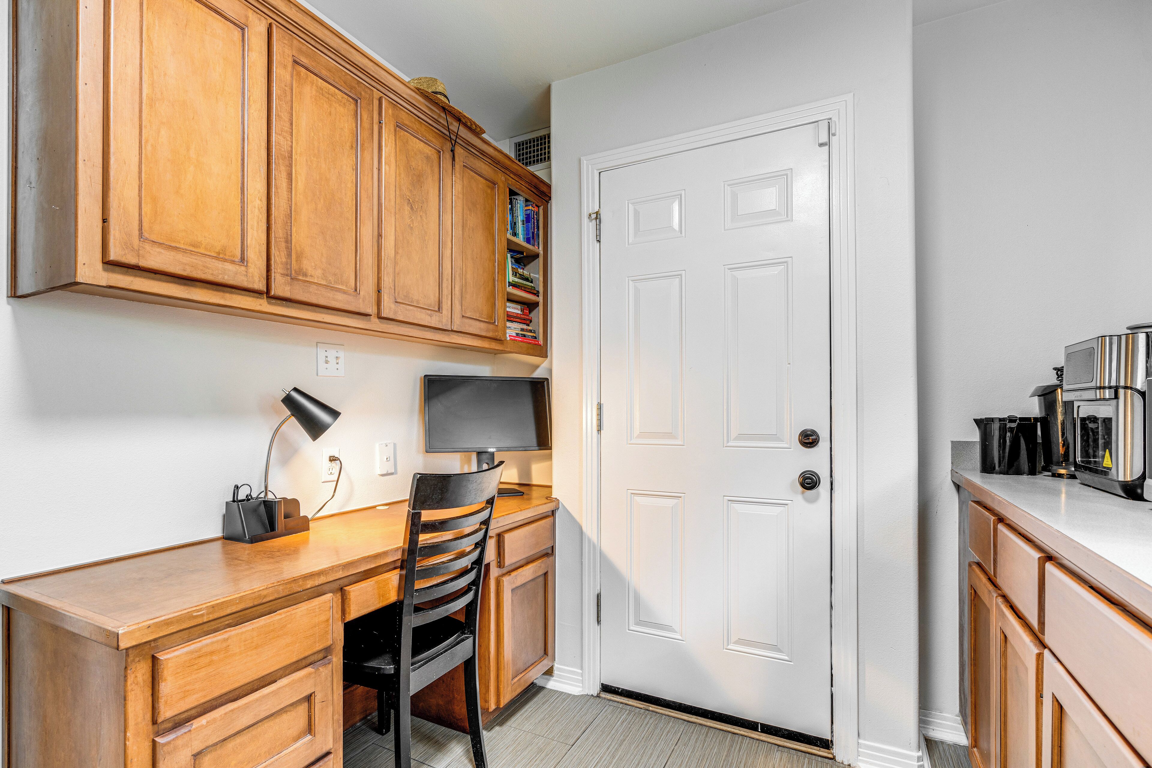 A compact kitchen with wooden cabinets, a built-in bookshelf, a desk with a computer, and a coffee maker.