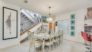 A dining area with a glass-top table and white chairs, a staircase with metal railings, and a chandelier.