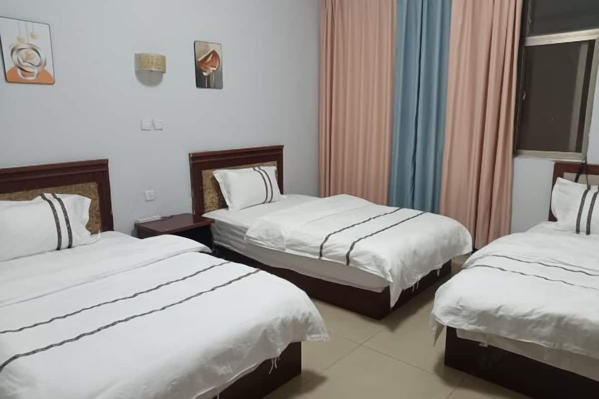 A hotel room with three single beds, a nightstand, a wall-mounted lamp, and a window with curtains.