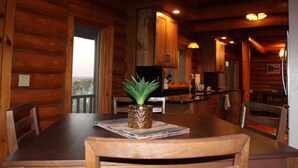 Interior de una cabaña de madera con una mesa de comedor, sillas, una pequeña planta en maceta y vistas a través de una ventana.