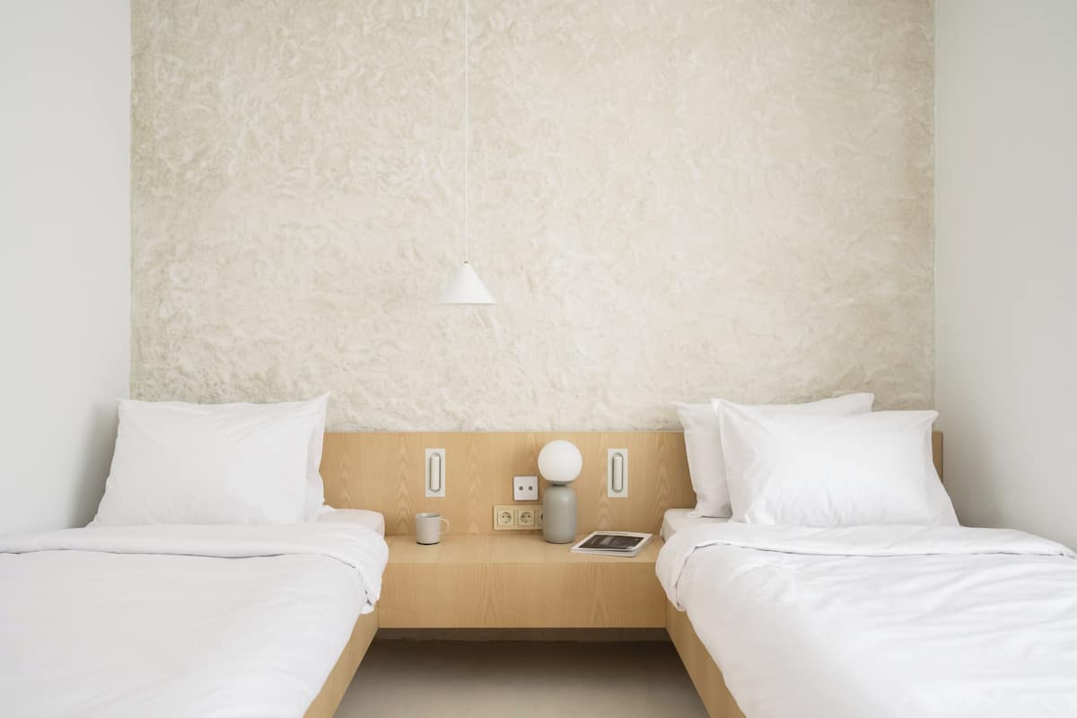 A minimalist hotel room with two single beds, a wooden headboard, a bedside table with a lamp, a cup, and a book.