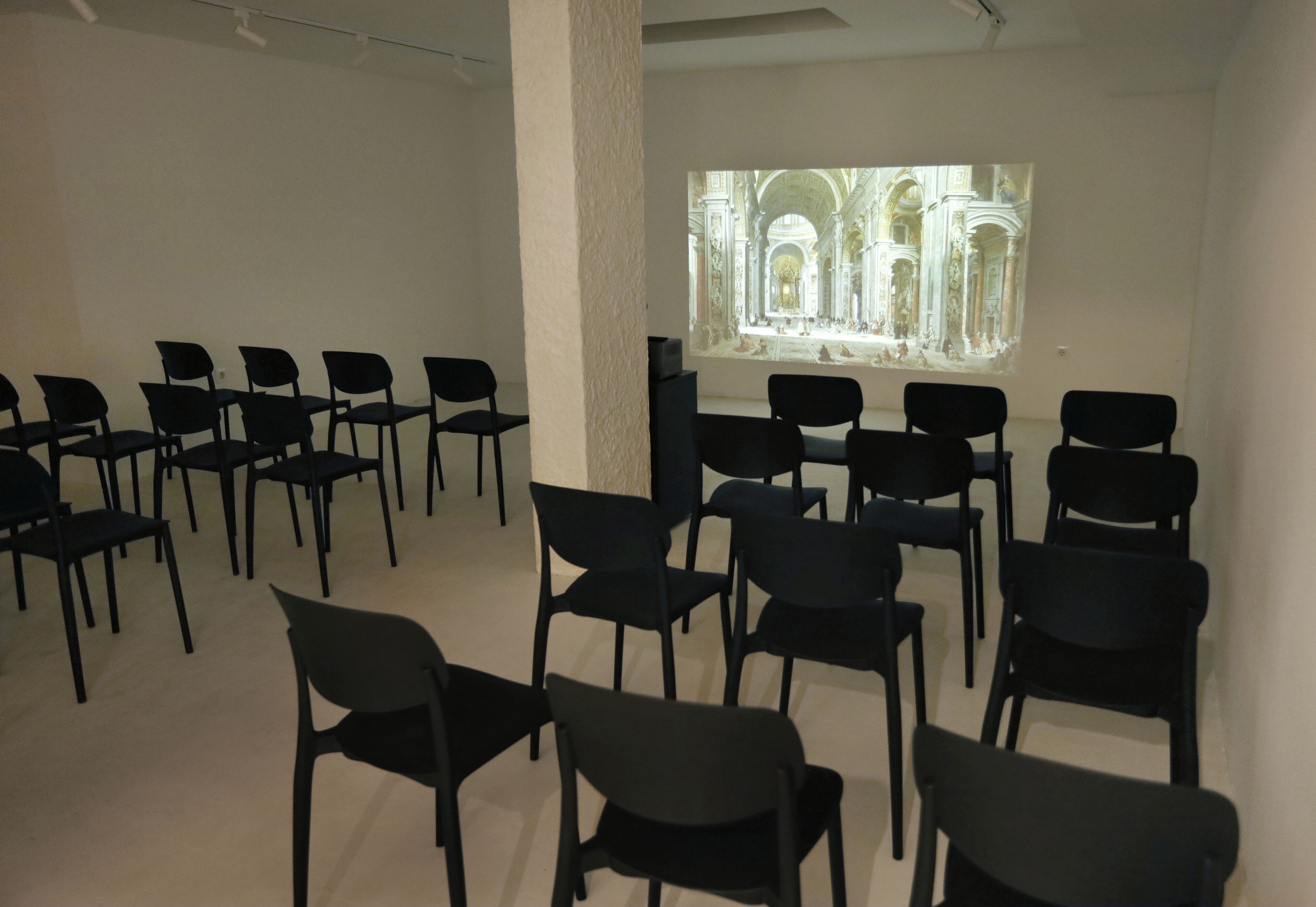 A room with rows of black chairs facing a large projection screen displaying an architectural image.
