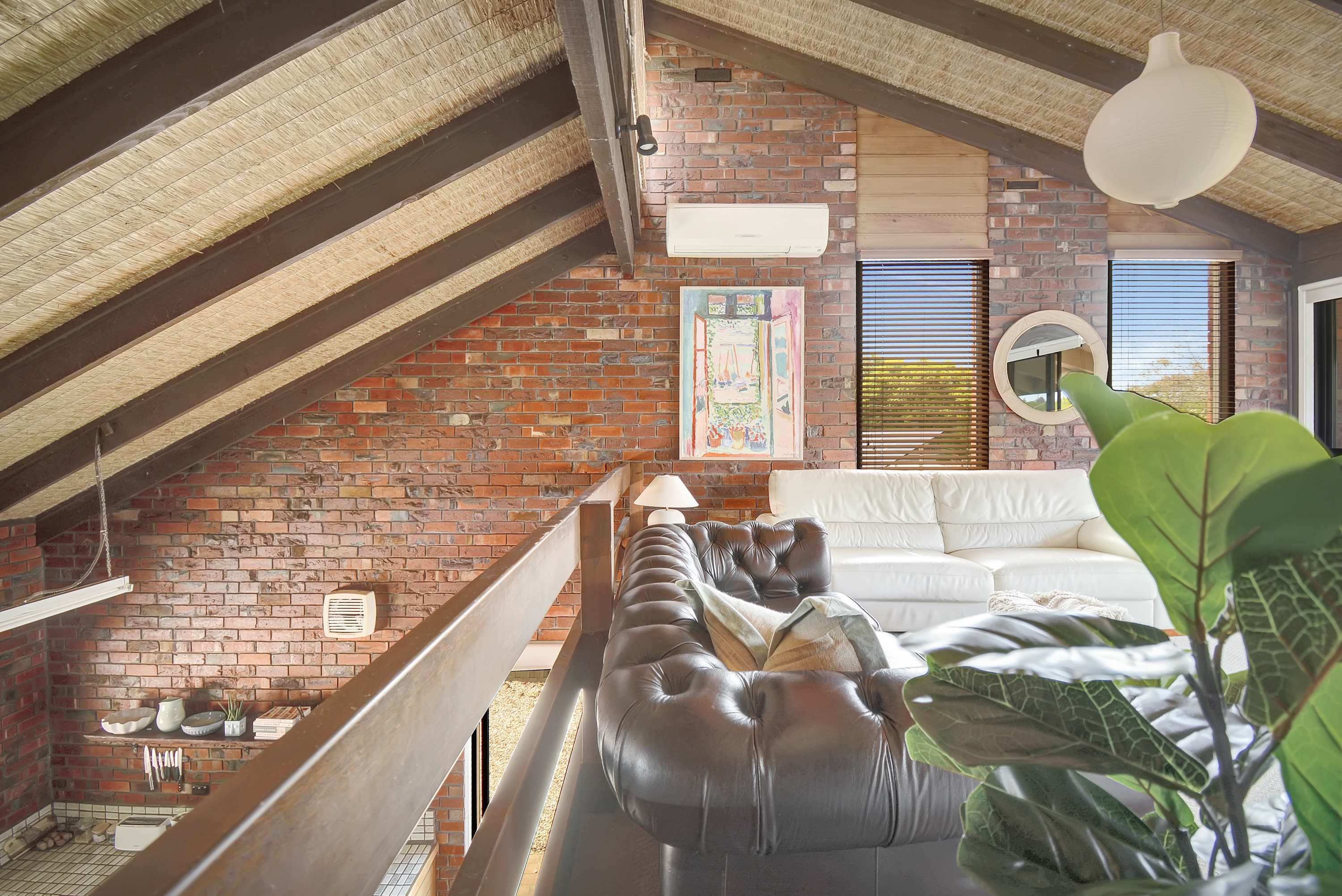 A loft-style room with exposed brick walls, a leather sofa, a hanging light fixture, and a large window with blinds.