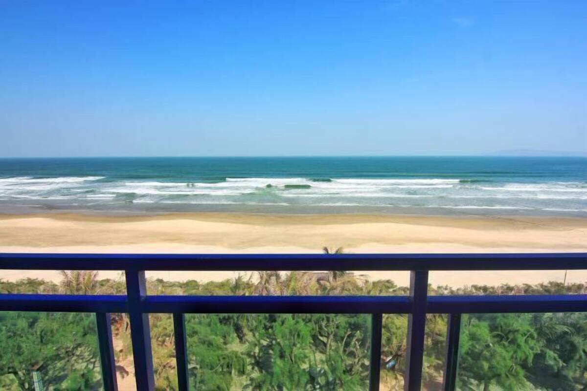 Une vue sur la plage depuis un balcon doté d’une balustrade, avec les vagues de l’océan et une végétation luxuriante au premier plan.