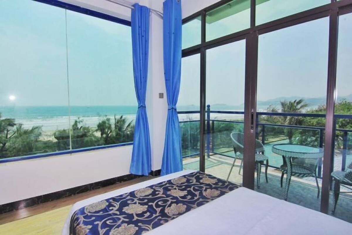 Une chambre avec un lit, une grande fenêtre avec des rideaux, un balcon avec une table et des chaises, et une vue sur la plage.