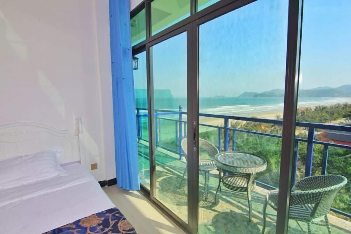 Une chambre avec un lit, un balcon offrant une vue sur la plage et les montagnes, et deux fauteuils en osier.