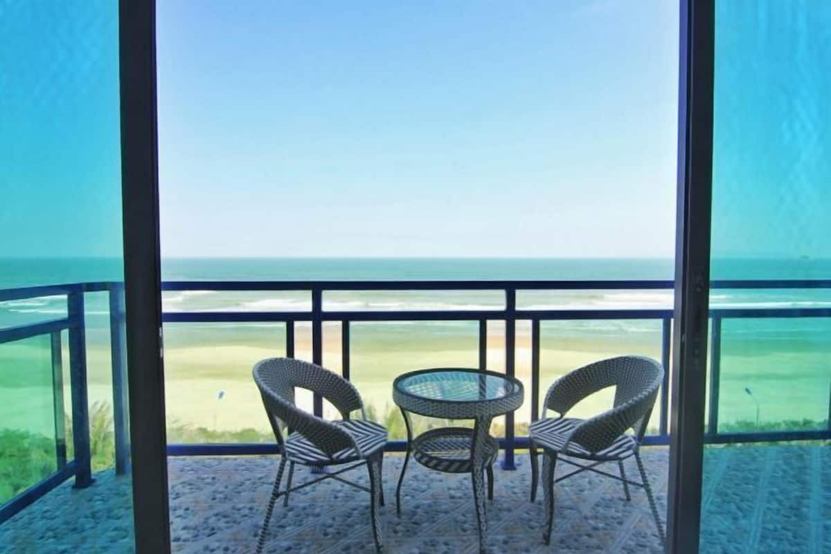 Un balcon avec des fauteuils en osier et une petite table, donnant sur une plage et l’océan.