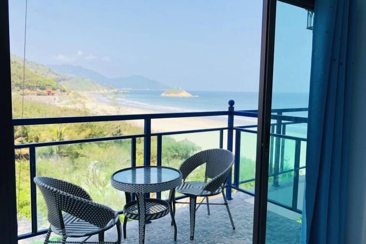 Un balcon avec deux fauteuils en osier et une table en verre, d’où l’on peut admirer une plage et des montagnes.