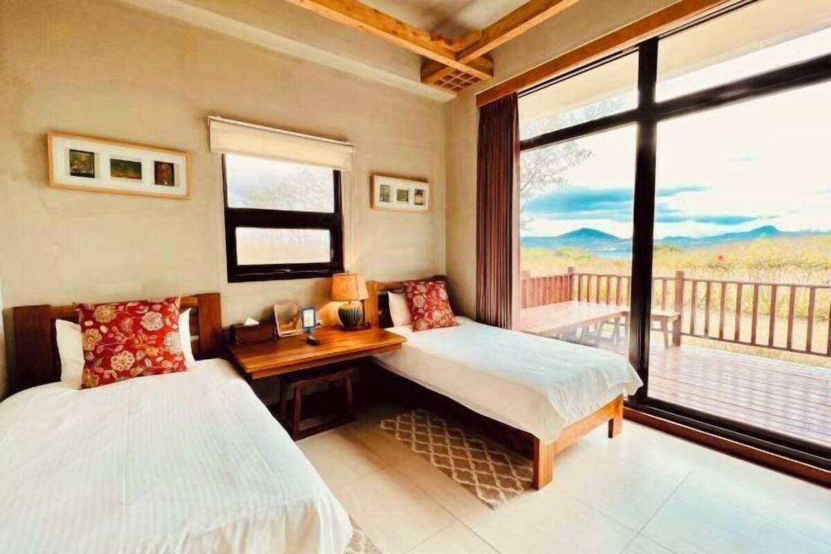 A room with two beds, a wooden desk, a window with a view of a balcony, and framed pictures on the wall.