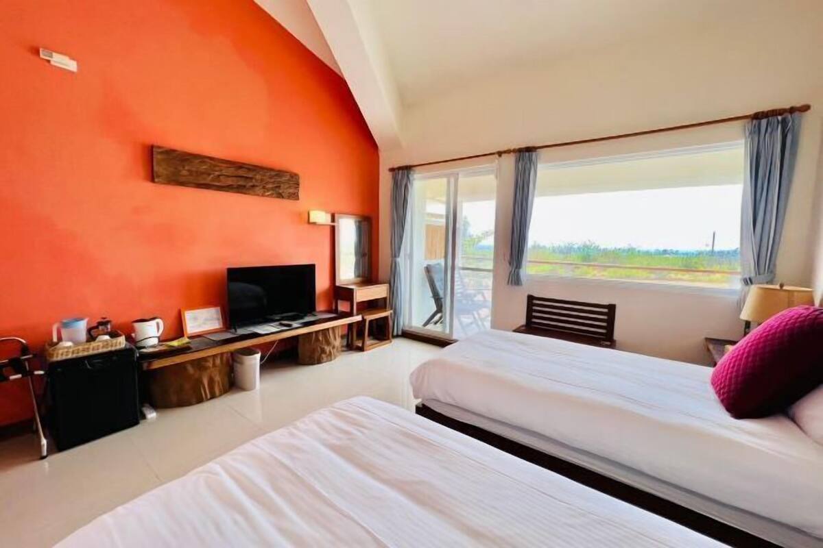A hotel room with two beds, a desk with a TV, a window with curtains, and a view of the outdoors.