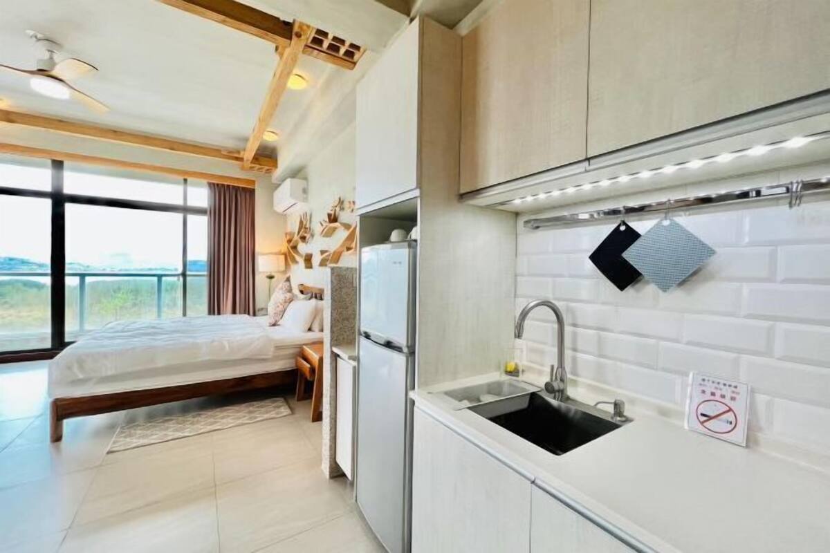 A modern bedroom with a large bed, a sitting area, and a view of the outdoors. The kitchen has white cabinets, a sink, and a no-smoking sign.
