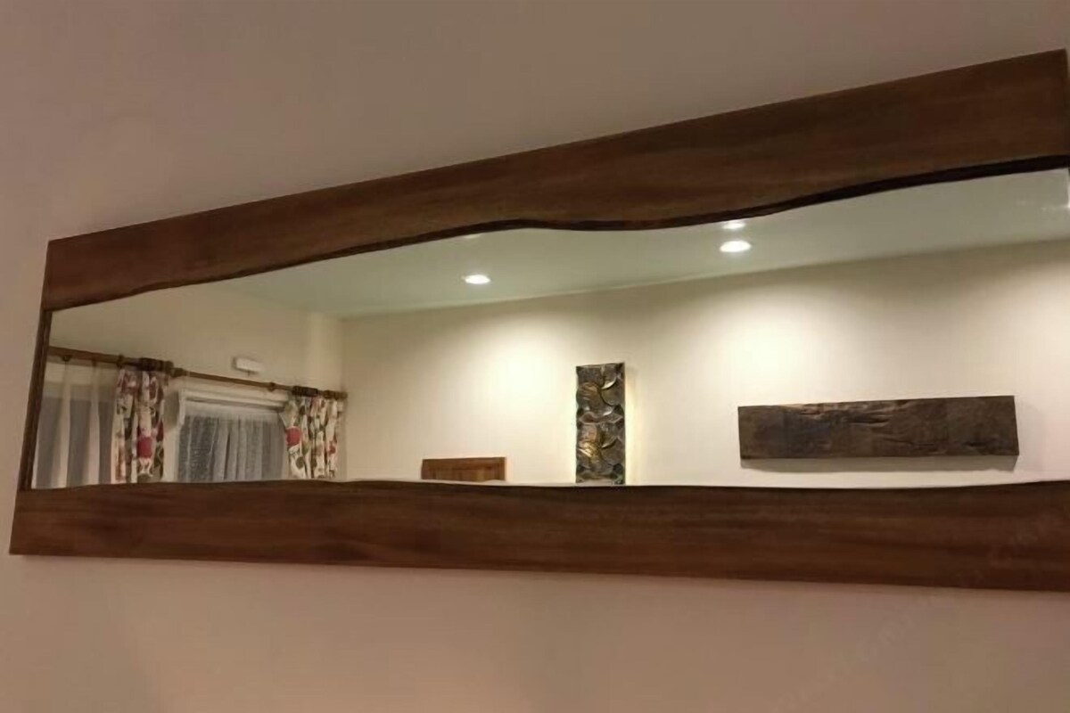 Un grand miroir avec un cadre en bois, une œuvre d’art accrochée au mur et une petite étagère sur laquelle se trouve un livre.