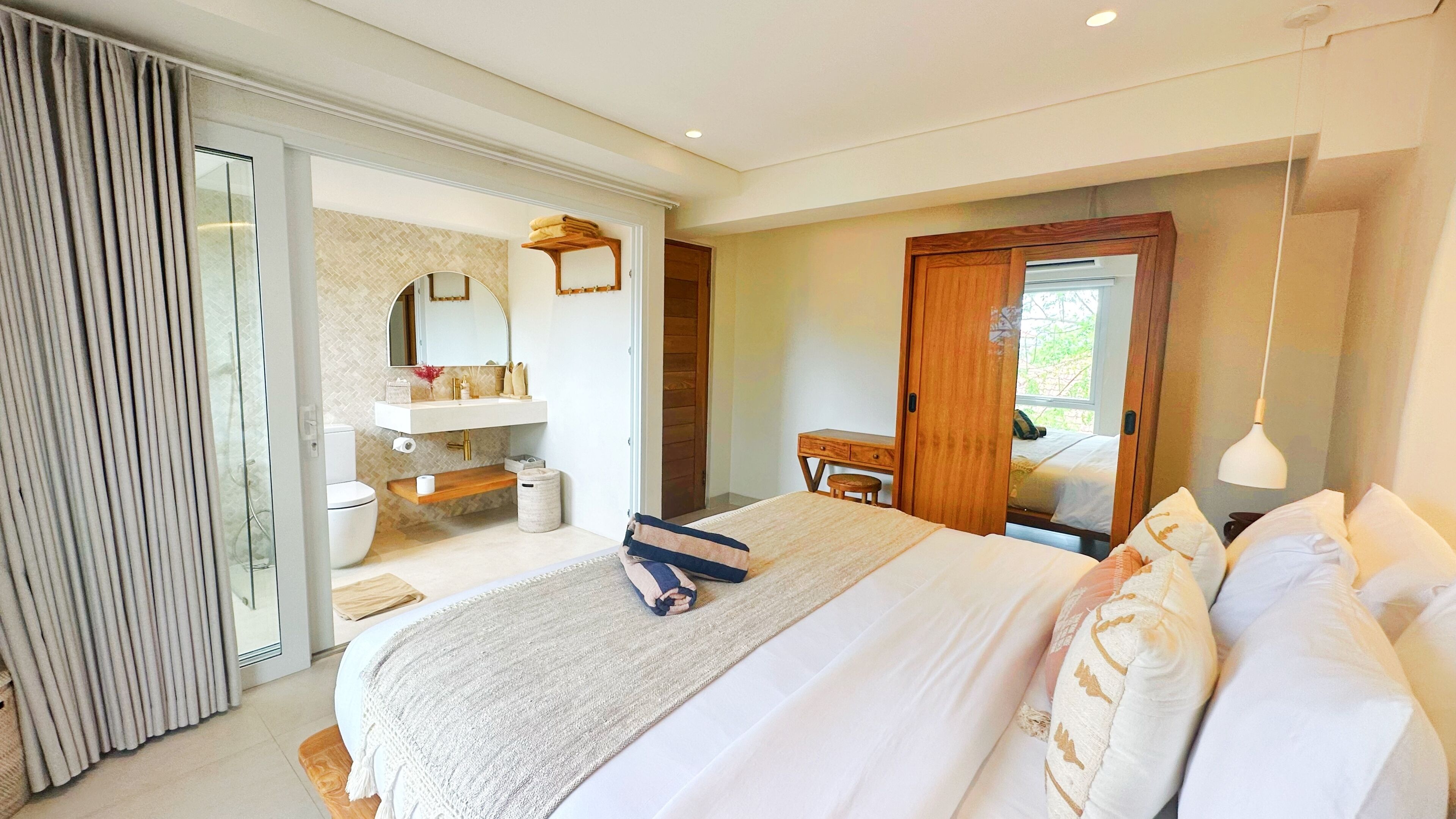 Ein Schlafzimmer mit einem großen Bett, einem Holztisch, einem Badezimmer mit Spiegel und Waschbecken sowie einer Schiebeglastür.