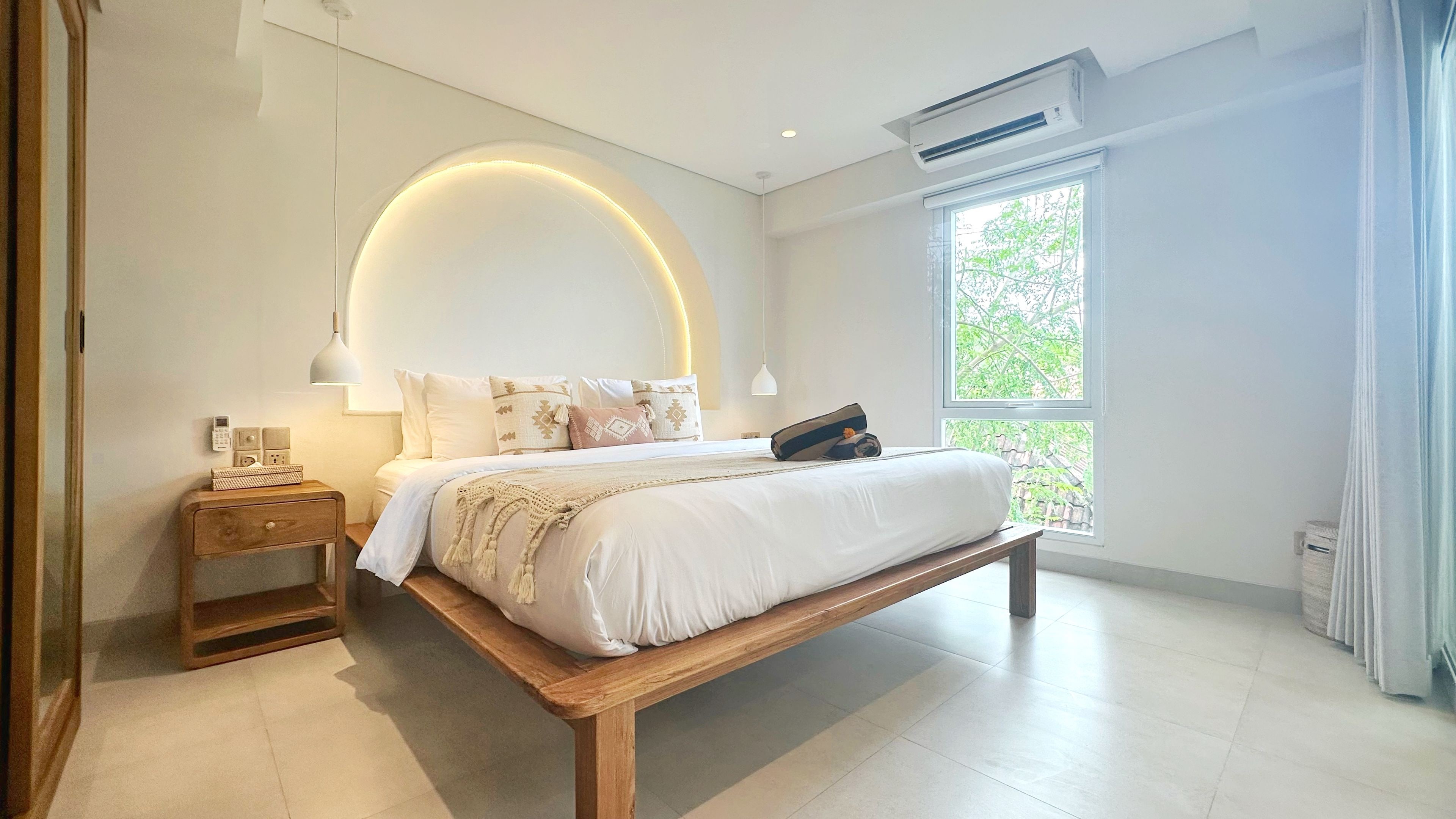 A modern bedroom with a large bed, bedside tables, a wall-mounted light, and a window with a view.