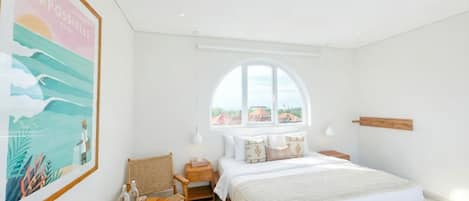 A bedroom with a large bed, a small table, a chair, a window with a view, and a wall-mounted shelf.